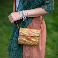 Person holding a woven handbag with a green cardigan and pink skirt against a blurred green background