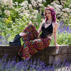 Woman with colorful pants and black top sitting on a wooden bench in a garden with flowers and lavender.