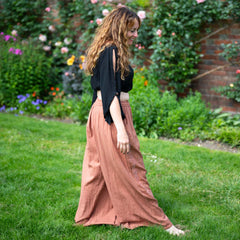 Woman in a black top and pink skirt standing in a garden with flowers and a brick wall in the background.
