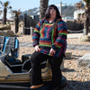 A plus size woman wearing a  Rainbow Baja Hoodie on a stony beach.