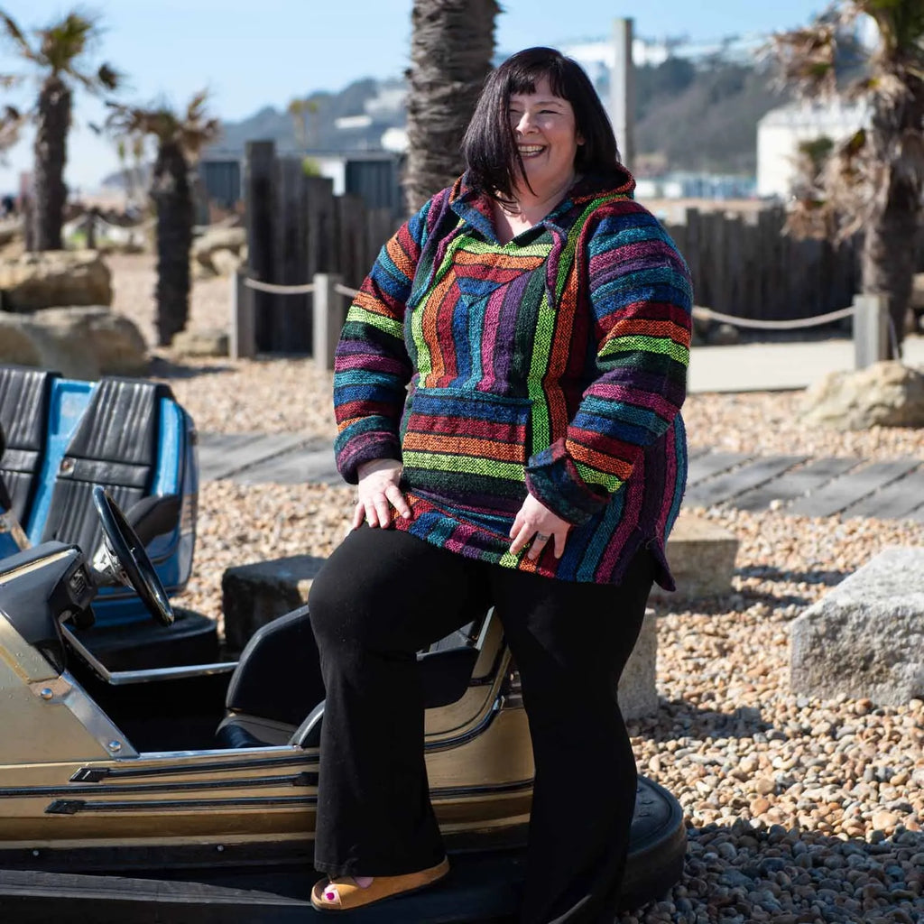 A plus size woman wearing a  Rainbow Baja Hoodie on a stony beach.