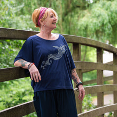 Woman wearing a blue shirt with a design, standing on a wooden bridge with greenery in the background.