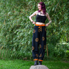 Woman wearing black top and high-waisted pants with sun patterns standing on a rock in a forest setting.