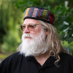 Man with a long white beard wearing a colorful patterned cap outdoors.