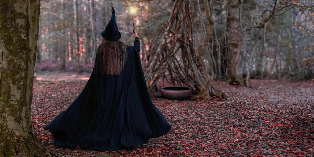 Person in a black witch's outfit walking through a forest with a wooden teepee in the background.